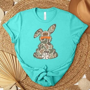 May include: A turquoise t-shirt featuring a cartoon bunny wearing a camouflage hat with the text "Eat Honey". The bunny is designed with a camouflage pattern and a white flower-shaped foot. The shirt is knotted at the bottom.