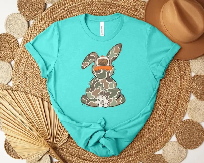 May include: A turquoise t-shirt featuring a cartoon bunny wearing a camouflage hat with the text "Eat Honey". The bunny is designed with a camouflage pattern and a white flower-shaped foot. The shirt is knotted at the bottom.