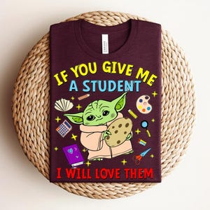 May include: Burgundy t-shirt with a graphic of a green alien-like character holding a cookie, surrounded by school supplies. Text reads "IF YOU GIVE ME A STUDENT I WILL LOVE THEM" in yellow and red.