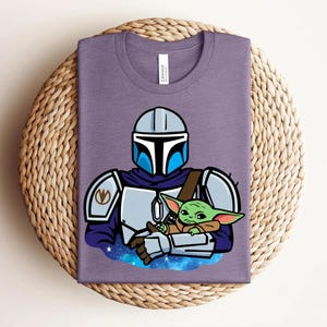 May include: A purple t-shirt featuring a cartoon illustration of a Mandalorian character holding a green alien. The Mandalorian is wearing silver armor with blue accents. The alien is wrapped in a brown blanket. The shirt has a crew neck.