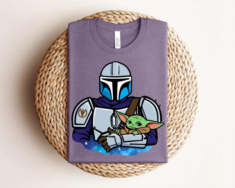 May include: A purple t-shirt featuring a cartoon illustration of a Mandalorian character holding a green alien. The Mandalorian is wearing silver armor with blue accents. The alien is wrapped in a brown blanket. The shirt has a crew neck.