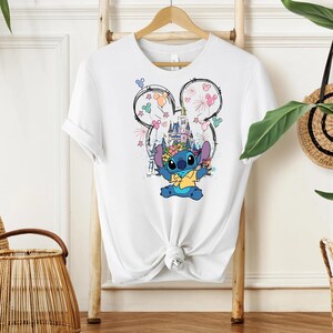 Pu&ograve; includere: T-shirt bianco con una grafica colorata di Stitch di Lilo & Stitch. Il design include un castello, fuochi d'artificio e un contorno della testa di Mickey Mouse. Stitch indossa una corona di fiori e una camicia gialla.