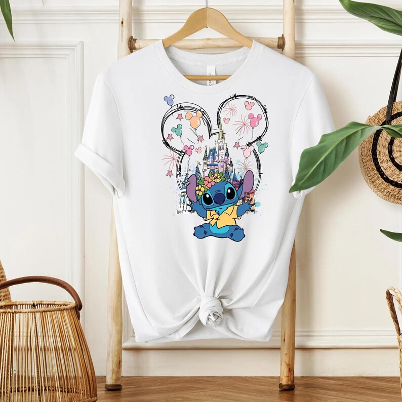 Pu&ograve; includere: T-shirt bianco con una grafica colorata di Stitch di Lilo & Stitch. Il design include un castello, fuochi d'artificio e un contorno della testa di Mickey Mouse. Stitch indossa una corona di fiori e una camicia gialla.