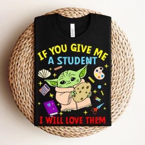 May include: Black t-shirt with a graphic of a green alien-like character holding a cookie, surrounded by school supplies. Text reads "IF YOU GIVE ME A STUDENT I WILL LOVE THEM" in yellow and red.