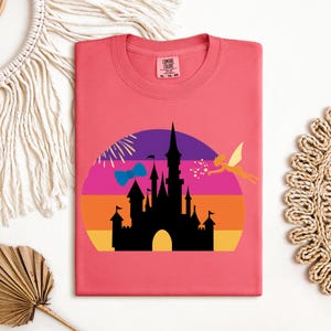 May include: A coral-colored t-shirt featuring a silhouette of a castle against a sunset backdrop. A fairy and fireworks are also included in the design. The shirt has a round neckline and a tag in the upper right corner.