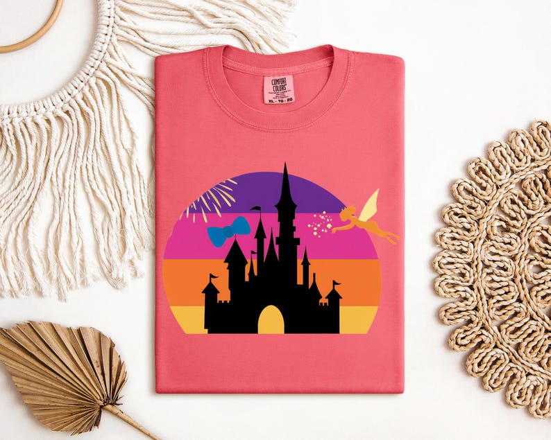May include: A coral-colored t-shirt featuring a silhouette of a castle against a sunset backdrop. A fairy and fireworks are also included in the design. The shirt has a round neckline and a tag in the upper right corner.