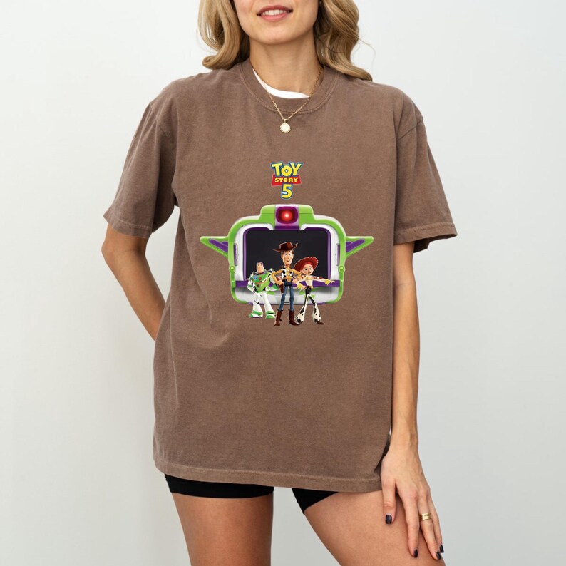 K&ouml;nnte beinhalten: Braunes T-Shirt mit einer Grafik von Toy Story 5-Charakteren in einem gr&uuml;n-wei&szlig;en Rahmen. Der Text "TOY STORY 5" befindet sich &uuml;ber dem Rahmen. Das T-Shirt wird von einer Person getragen.