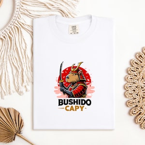 May include: White t-shirt featuring a cartoon capybara in samurai armor holding a sword against a red sun design. The text "BUSHIDO - CAPY" is printed below the graphic. The shirt is made of a soft material.
