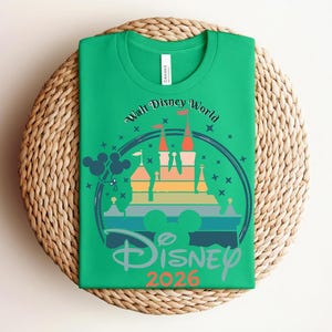 Op de afbeelding: Groen t-shirt met een kleurrijke afbeelding van een kasteel, Mickey Mouse oren en de tekst "Walt Disney World" en "Disney 2026". Het shirt is opgevouwen op een ronde, geweven placemat.