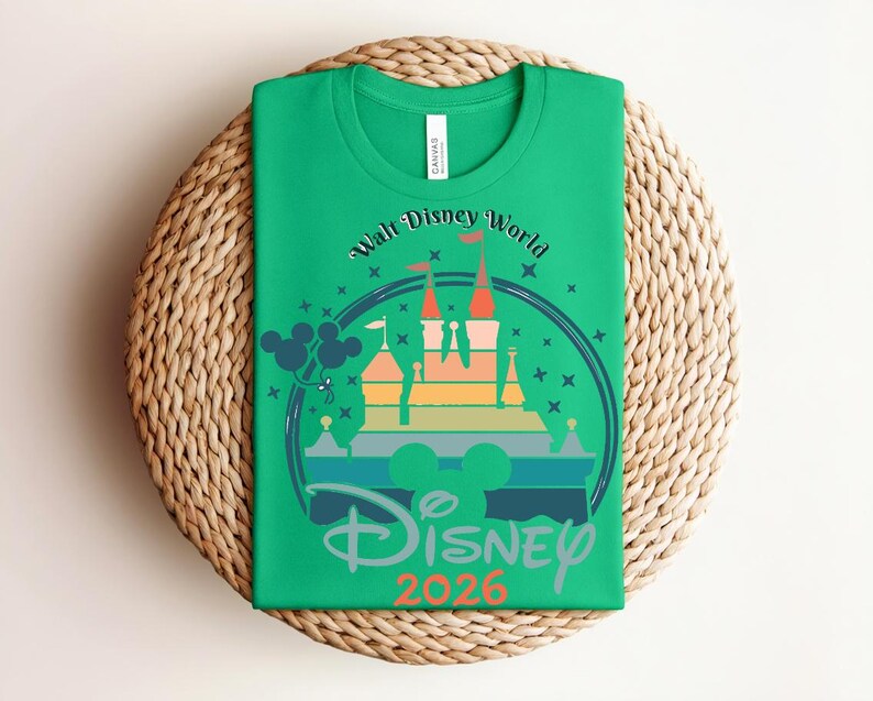 Op de afbeelding: Groen t-shirt met een kleurrijke afbeelding van een kasteel, Mickey Mouse oren en de tekst "Walt Disney World" en "Disney 2026". Het shirt is opgevouwen op een ronde, geweven placemat.