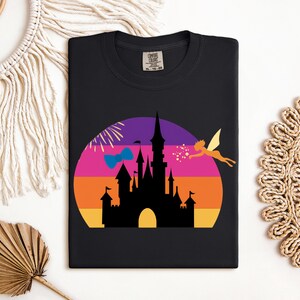 May include: Black t-shirt featuring a colorful graphic of a castle silhouette against a sunset backdrop. A fairy and fireworks are also included in the design. The shirt is a Comfort Colors brand.