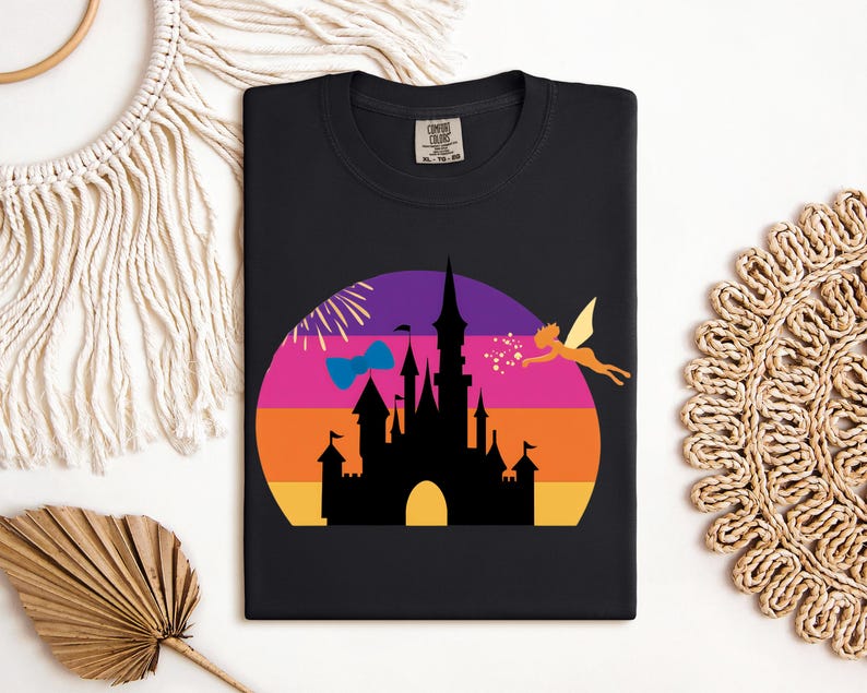 May include: Black t-shirt featuring a colorful graphic of a castle silhouette against a sunset backdrop. A fairy and fireworks are also included in the design. The shirt is a Comfort Colors brand.