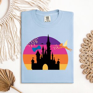May include: Light blue t-shirt featuring a colorful graphic of a castle silhouette against a sunset backdrop. A fairy and fireworks are also included in the design. The shirt has a crew neck and a tag on the collar.