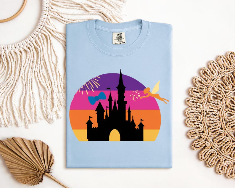 May include: Light blue t-shirt featuring a colorful graphic of a castle silhouette against a sunset backdrop. A fairy and fireworks are also included in the design. The shirt has a crew neck and a tag on the collar.