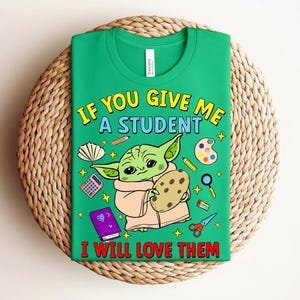 May include: Green t-shirt with the text "IF YOU GIVE ME A STUDENT I WILL LOVE THEM" and a cartoon character holding a cookie. The shirt has school-themed graphics like a calculator, paint palette, and scissors.