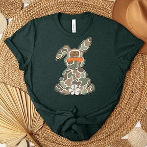 May include: Dark green t-shirt featuring a camouflage bunny graphic wearing an orange hat. The bunny has a white flower on its chest. The shirt is displayed on a woven mat with a brown hat in the background.