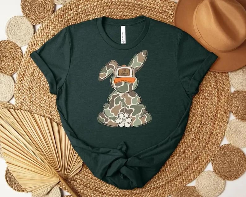 May include: Dark green t-shirt featuring a camouflage bunny graphic wearing an orange hat. The bunny has a white flower on its chest. The shirt is displayed on a woven mat with a brown hat in the background.