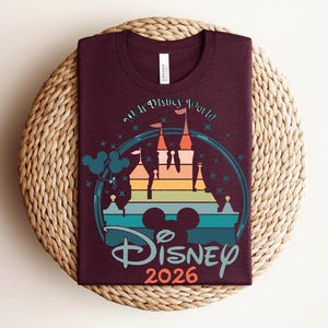 Op de afbeelding: Bordeauxkleurig T-shirt met een kleurrijke afbeelding van een kasteel, Mickey Mouse oren en de tekst "Disney 2026". Het ontwerp heeft een regenboogkleurig kasteelsilhouet met turquoise, oranje en blauwe accenten. Het shirt wordt gepresenteerd op een geweven, cirkelvormig oppervlak.