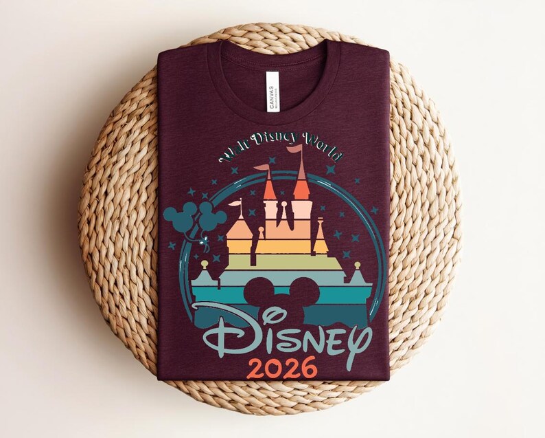 Op de afbeelding: Bordeauxkleurig T-shirt met een kleurrijke afbeelding van een kasteel, Mickey Mouse oren en de tekst "Disney 2026". Het ontwerp heeft een regenboogkleurig kasteelsilhouet met turquoise, oranje en blauwe accenten. Het shirt wordt gepresenteerd op een geweven, cirkelvormig oppervlak.