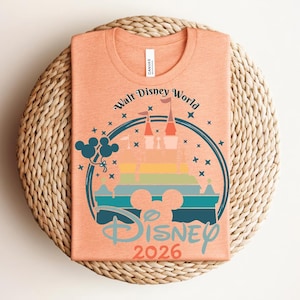 Op de afbeelding: Perzikkleurig T-shirt met een kleurrijke afbeelding van een kasteel, Mickey Mouse-silhouet en ballonnen. De tekst luidt "Walt Disney World" en "Disney 2026" in een cirkelvormig ontwerp. Het shirt is op een geweven mat gevouwen.