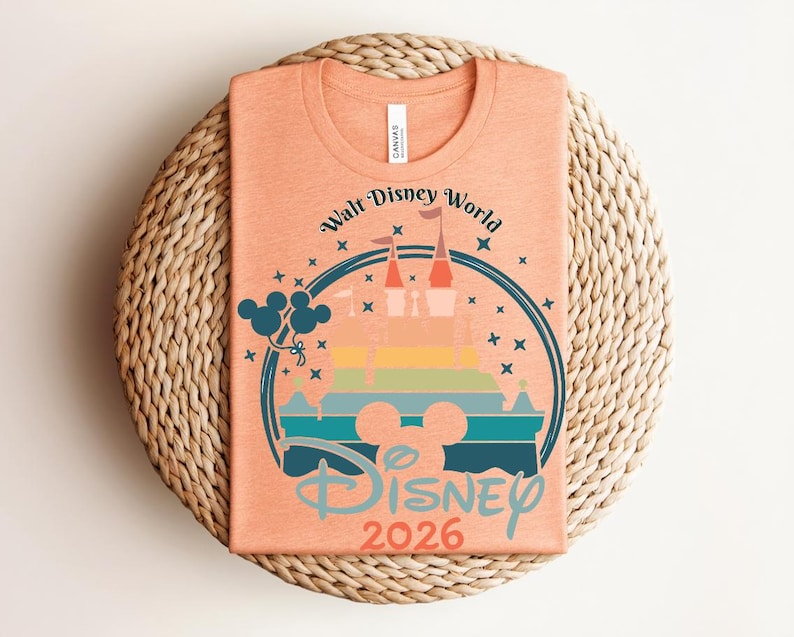 Op de afbeelding: Perzikkleurig T-shirt met een kleurrijke afbeelding van een kasteel, Mickey Mouse-silhouet en ballonnen. De tekst luidt "Walt Disney World" en "Disney 2026" in een cirkelvormig ontwerp. Het shirt is op een geweven mat gevouwen.