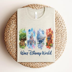 May include: A light beige t-shirt featuring a watercolor design of Walt Disney World landmarks. The design includes the Tree of Life, Cinderella Castle, Spaceship Earth, and the Tower of Terror, with the words "Walt Disney World" in blue.