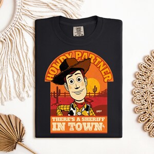 K&ouml;nnte beinhalten: Schwarzes T-Shirt mit einer Cartoon-Illustration von Woody aus Toy Story. Das Design enth&auml;lt den Text "HOWDY PARTNER" &uuml;ber Woodys Bild und "THERE'S A SHERIFF IN TOWN" darunter. Das T-Shirt hat einen klassischen Rundhalsausschnitt.