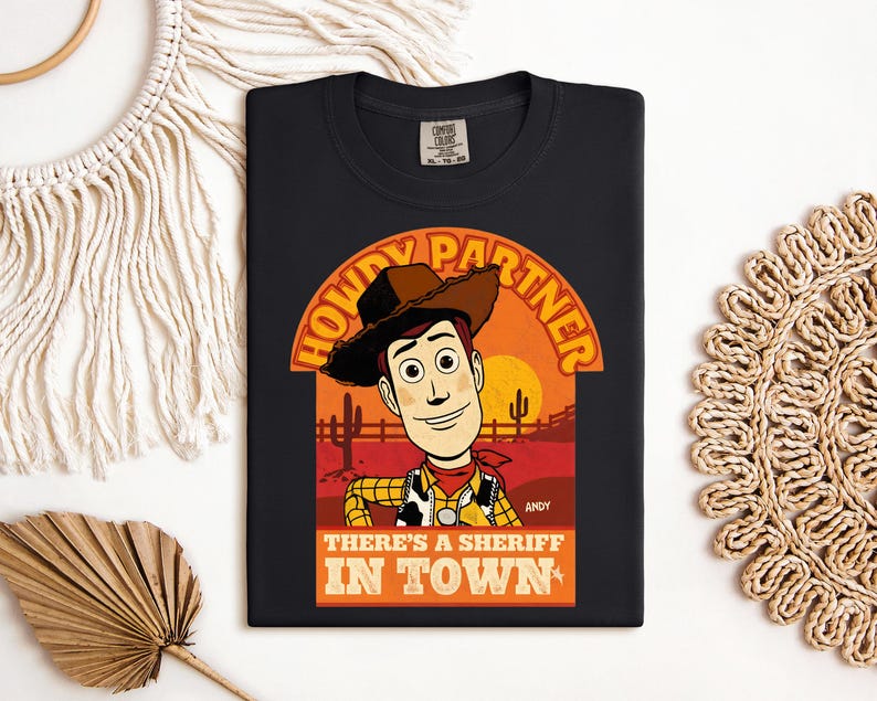 K&ouml;nnte beinhalten: Schwarzes T-Shirt mit einer Cartoon-Illustration von Woody aus Toy Story. Das Design enth&auml;lt den Text "HOWDY PARTNER" &uuml;ber Woodys Bild und "THERE'S A SHERIFF IN TOWN" darunter. Das T-Shirt hat einen klassischen Rundhalsausschnitt.
