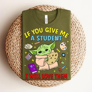 May include: Olive green t-shirt featuring a cartoon character holding a cookie, surrounded by school-themed illustrations. The text on the shirt reads, "IF YOU GIVE ME A STUDENT I WILL LOVE THEM."