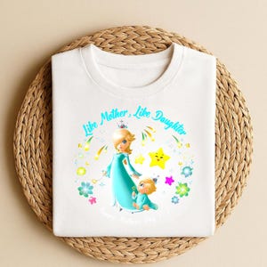 Puede incluir: Camiseta blanca con una ilustraci&oacute;n de dibujos animados de una madre y un ni&ntilde;o con vestidos turquesa, con el texto "Like Mother, Like Daughter" y "Happy Mother's Day 2023". El dise&ntilde;o incluye estrellas y flores.