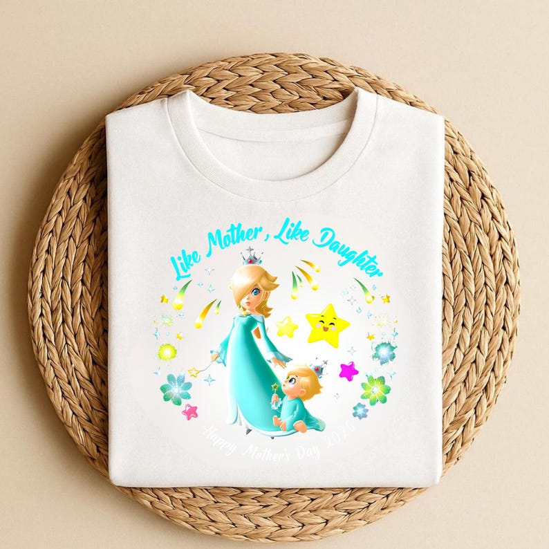Puede incluir: Camiseta blanca con una ilustraci&oacute;n de dibujos animados de una madre y un ni&ntilde;o con vestidos turquesa, con el texto "Like Mother, Like Daughter" y "Happy Mother's Day 2023". El dise&ntilde;o incluye estrellas y flores.