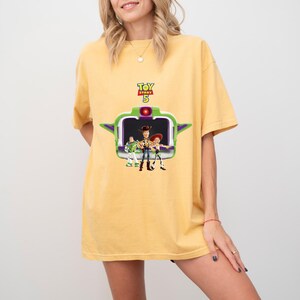K&ouml;nnte beinhalten: Gelbes T-Shirt mit einer Grafik aus dem Film Toy Story 5. Die Grafik zeigt den Filmtitel und die Charaktere Woody, Buzz Lightyear und Jessie. Das Shirt ist einfarbig und hat kurze &Auml;rmel.