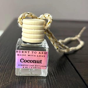 May include: A small square glass car diffuser bottle with a light wood cap and a braided beige and gold cord. The pink label reads: BURNT TO ASH, MADE WITH LOVE, Coconut, SCENTED CAR DIFFUSER, 8 ML / 8 GRAMS. The bottle is clear and empty.