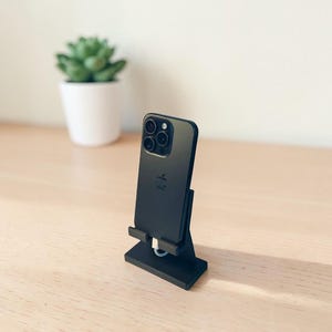 May include: A black smartphone is propped up on a black stand, with a white charging cable plugged in. The phone has a triple-lens camera. A small succulent plant in a white pot sits in the background on a light wood surface.