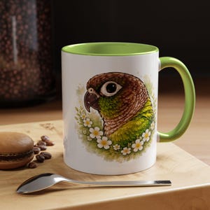 Könnte beinhalten: Weiße Keramik-Tasse mit grünem Innenraum und Griff. Die Tasse zeigt eine detaillierte Illustration eines Grünwangen-Sittichs, umgeben von weißen Blumen. Ein Löffel und ein Gebäck liegen auf einer Holzoberfläche.
