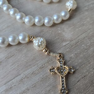 May include: A rosary with large, round, off-white beads and gold-colored accents. The rosary features a gold-colored cross pendant adorned with small, clear crystals. The beads are strung together, creating a religious accessory.