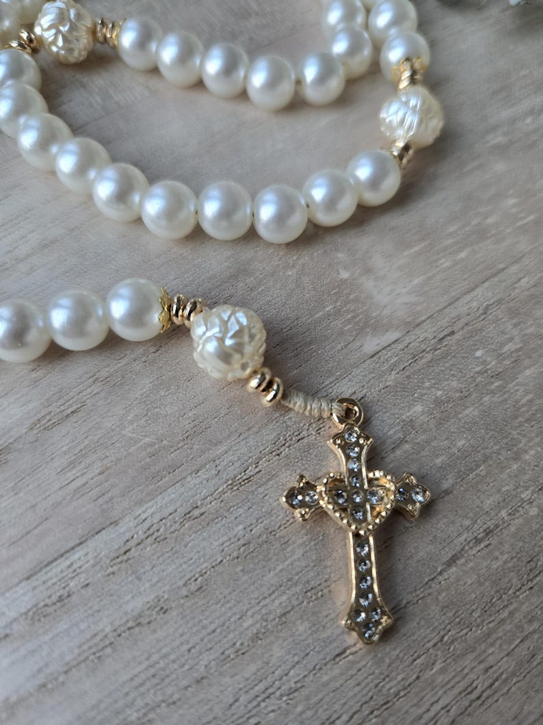 May include: A rosary with large, round, off-white beads and gold-colored accents. The rosary features a gold-colored cross pendant adorned with small, clear crystals. The beads are strung together, creating a religious accessory.