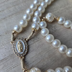 May include: A rosary featuring large, creamy white faux pearls. The centerpiece has a gold-toned oval pendant with a religious figure and is surrounded by small, clear crystals. Gold-toned accents are present throughout the rosary.