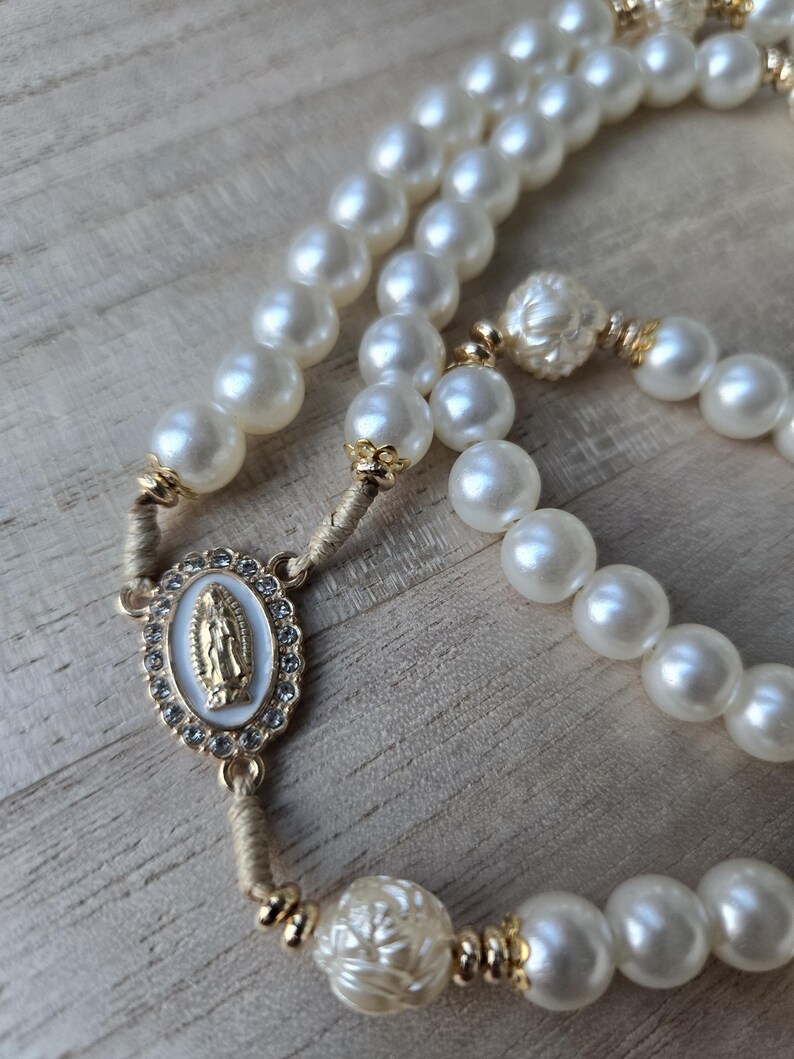 May include: A rosary featuring large, creamy white faux pearls. The centerpiece has a gold-toned oval pendant with a religious figure and is surrounded by small, clear crystals. Gold-toned accents are present throughout the rosary.