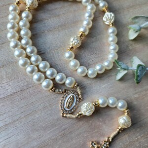 May include: A pearl rosary with gold accents and a decorative cross. The rosary features large, round, off-white pearls, gold-colored connectors, and a gold-toned cross with clear crystal accents. The centerpiece has an image of the Virgin Mary.