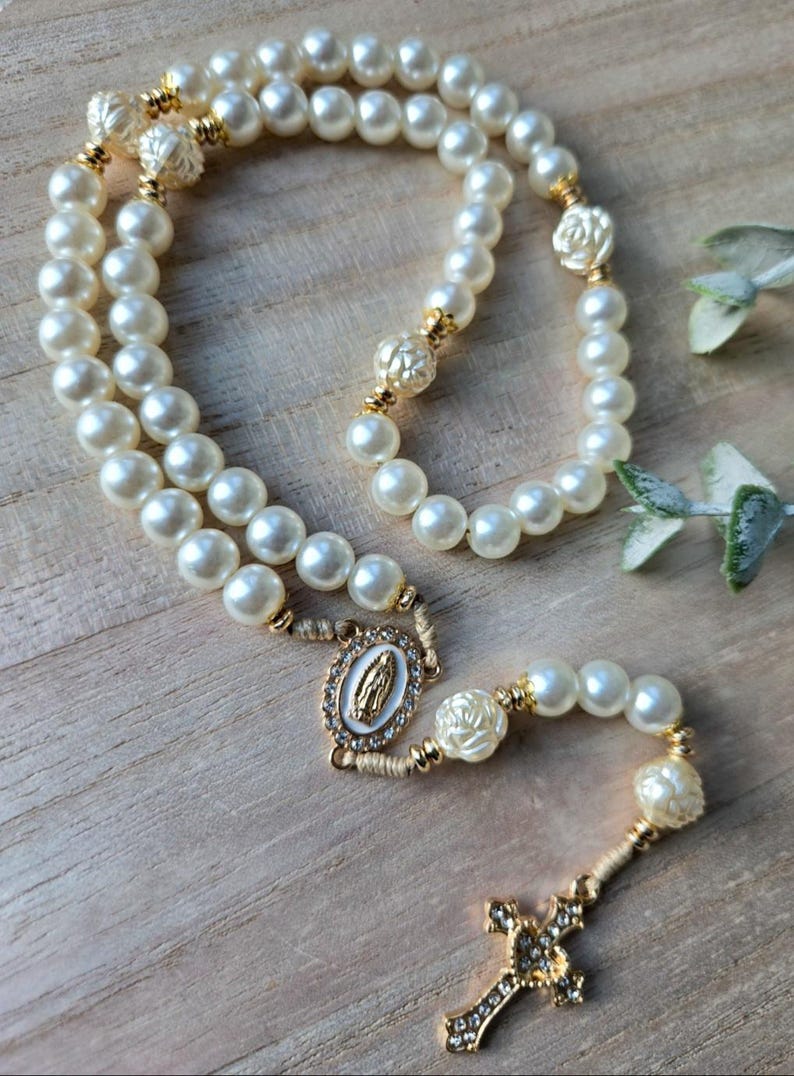 May include: A pearl rosary with gold accents and a decorative cross. The rosary features large, round, off-white pearls, gold-colored connectors, and a gold-toned cross with clear crystal accents. The centerpiece has an image of the Virgin Mary.