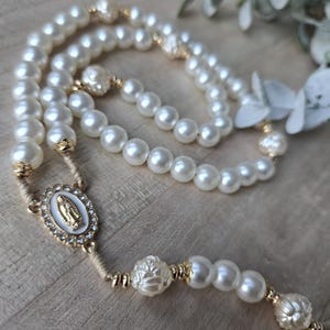 May include: A pearl rosary with gold accents. The rosary features large, round, off-white pearls and decorative gold-colored beads. An oval centerpiece with a religious image is also present.