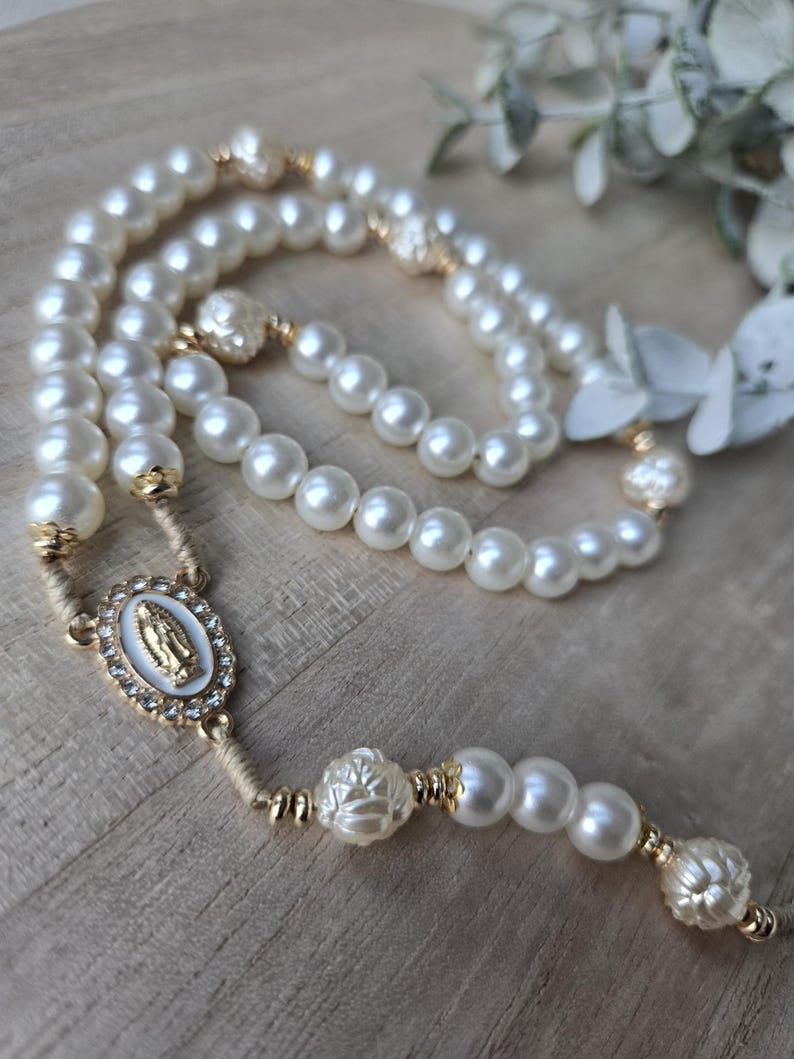 May include: A pearl rosary with gold accents. The rosary features large, round, off-white pearls and decorative gold-colored beads. An oval centerpiece with a religious image is also present.