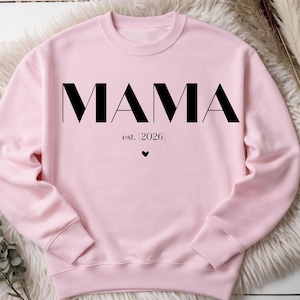 May include: A light pink sweatshirt with the word "MAMA" in large, black, bold letters. Below the word "MAMA" is the text "est. [2026]" and a small black heart. The sweatshirt is made of a soft material.
