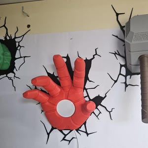 Può includere: Un pugno verde, una mano rossa con un cerchio bianco e un martello grigio con manico marrone sono montati su un muro bianco. Decalcomanie di crepe nere circondano ogni oggetto, creando un effetto 3D. Gli oggetti sono a tema personaggi dei fumetti.