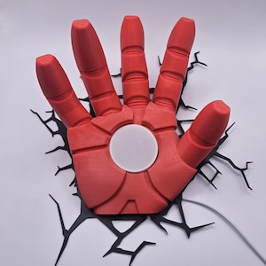 Op de afbeelding: Een rode, 3D-geprinte Iron Man-hand met een wit cirkelvormig midden, gemonteerd op een zwart, scheurachtig ontwerp. De hand is gedetailleerd met gesegmenteerde vingers en een getextureerd oppervlak. Een wit snoer loopt vanaf de basis.