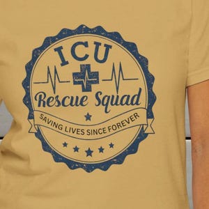 Può includere: T-shirt giallo senape con un emblema blu navy. L'emblema recita "ICU Rescue Squad" e presenta un simbolo a croce. È incluso anche il testo "Saving Lives Since Forever". La maglietta è realizzata in un materiale morbido.