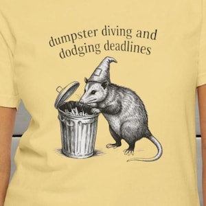 Op de afbeelding: Lichtgeel T-shirt met een zwart-wit illustratie van een opossum met een tovenaars hoed naast een vuilnisbak. De tekst "dumpster diving and dodging deadlines" staat boven de illustratie.