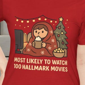 Könnte beinhalten: Rotes T-Shirt mit dem Aufdruck "MOST LIKELY TO WATCH 100 HALLMARK MOVIES". Die Grafik zeigt eine Person in einer Decke, die fernsieht, mit einem Weihnachtsbaum und Popcorn.