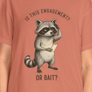 May include: Coral-colored t-shirt featuring a cartoon raccoon holding a paper and pondering. The text on the shirt reads "IS THIS ENGAGEMENT? OR BAIT?" The raccoon is drawn in shades of gray and white.
